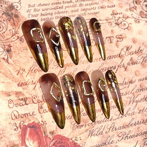 May include: Set of ten long, almond-shaped false nails with a brown and gold design. Each nail has a gold accent and a geometric design. The nails are on a background with a floral pattern and text that reads "Registered" and "Not a flower".