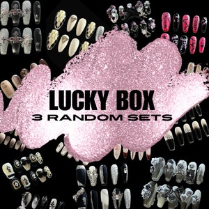May include: A pink glitter background with the text "LUCKY BOX 3 RANDOM SETS". The image features a variety of press-on nail sets with different designs, including black and white, glitter, and floral patterns.