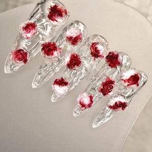 May include: Clear acrylic nail set with a pointed shape, each nail adorned with a 3D red and white rose design. The nails are arranged on a light gray surface, showcasing the intricate floral details.