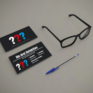 May include: Two black business cards with white question marks and the text "Die drei Detektive" in red, blue, and white. The text on the second card reads "Wer &uuml;berlebt jeden Fall?" and lists the detectives' names and roles. A blue pen and black-framed glasses are also on the table.