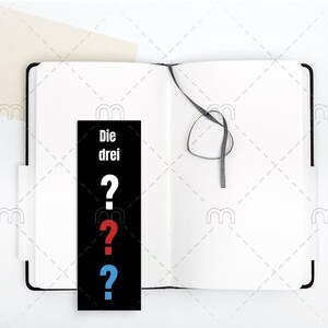 May include: A black and white bookmark with the text "Die drei" and three question marks in red, blue, and white. The bookmark is placed in a white notebook with a black ribbon.