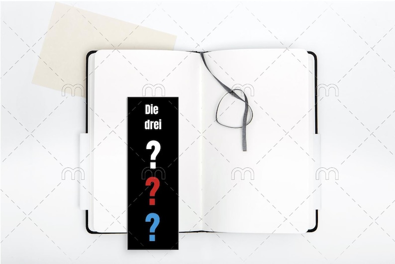 May include: A black and white bookmark with the text "Die drei" and three question marks in red, blue, and white. The bookmark is placed in a white notebook with a black ribbon.