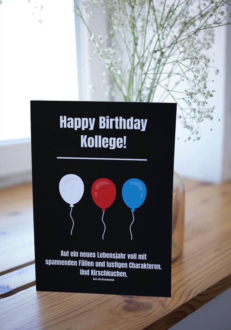May include: A black card with white text that says "Happy Birthday Kollege!" with three balloons in the middle, one white, one red, and one blue. The text below the balloons says "Auf ein neues Lebensjahr voll mit spannenden F&auml;llen und lustigen Charakteren. Und Kirschkuchen."
