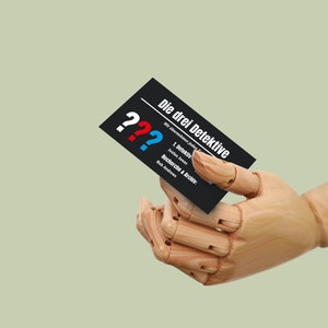 May include: A black business card with white text that reads "Die drei Detektive" and three question marks in red, blue, and white. The card is held by a wooden hand.