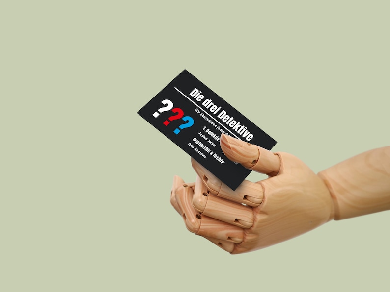 May include: A black business card with white text that reads "Die drei Detektive" and three question marks in red, blue, and white. The card is held by a wooden hand.