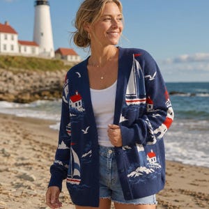 May include: A navy blue button-down cardigan featuring a repeating knit pattern of white sailboats, red and white lighthouses, and seagulls. The garment is worn over a plain white tank top and blue denim shorts, showcasing a nautical-themed design.