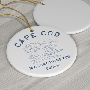 May include: White ceramic ornament with a blue outline of a lighthouse, houses, and clouds. The text "CAPE COD MASSACHUSETTS Estd. 1602" is printed in blue.