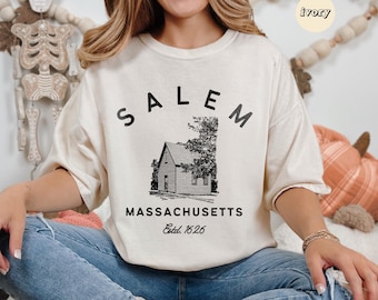 Könnte beinhalten: Ein cremefarbener Pullover mit einer schwarzen Grafik eines Hauses und dem Text "SALEM MASSACHUSETTS Est. 1626".