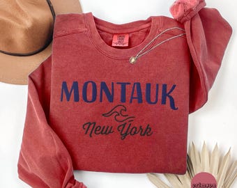May include: A red-orange sweatshirt with the word "MONTAUK" in navy blue block letters, with "New York" below. A gold necklace with a pendant is draped across the sweatshirt. A brown hat is visible in the background.
