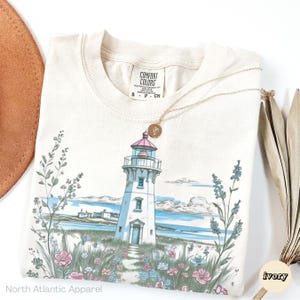 May include: Ivory-colored t-shirt featuring a lighthouse illustration with a pink roof, surrounded by flowers and greenery. A gold necklace with a pendant rests on the shirt. The text "North Atlantic Apparel" is visible at the bottom.