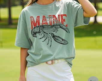 May include: Sage green t-shirt with the word "MAINE" in red block letters above a detailed black and white illustration of a lobster. The shirt is a comfortable, casual style.