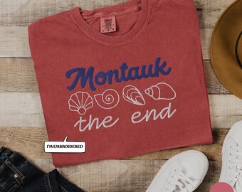 May include: A red t-shirt with the embroidered text "Montauk the end" in blue and white, along with seashell graphics. The shirt is paired with white sneakers, a brown hat, and denim jeans on a wooden surface.