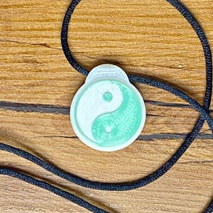 以下が含まれることがあります： 黒い紐のネックレスに、白と緑の陰陽のシンボルペンダントが付いている。