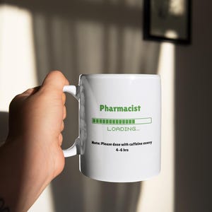 May include: White ceramic mug with the word "Pharmacist" in green, a loading bar graphic, and the text "LOADING...". Below, it reads "Note: Please dose with caffeine every 4-6 hrs".