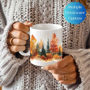 May include: White ceramic mug with a watercolor design of an autumn forest scene. The trees are in shades of orange, yellow, and green. The mug is being held by a hand.