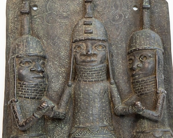 Pode incluir: Escultura em relevo de bronze retratando três figuras com chapéus e vestimentas ornamentadas. A figura central é ladeada por outras duas, todas segurando objetos. O fundo apresenta padrões florais e uma superfície texturizada.