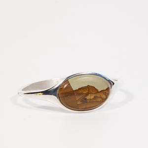 Puede incluir: Un brazalete plateado con una piedra ovalada en el centro. La piedra presenta un diseño de paisaje en tonos marrón, beige y crema. El brazalete tiene un acabado pulido. La piedra mide unos 3 cm de largo.