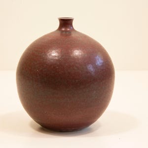 Könnte beinhalten: Eine runde, weinrote Keramikvase mit schmalem Hals. Die Vase hat eine strukturierte Oberfläche und ein glänzendes Finish. Die Form erinnert an einen Granatapfel. Die Vase ist ein dekorativer Gegenstand.