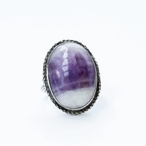 May include: A silver ring featuring a large oval amethyst gemstone. The amethyst is a deep purple colour with white streaks.