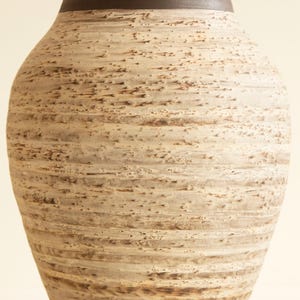 May include: A tall, textured vase with a wide, rounded body and a dark brown rim. The vase has a light beige and brown color scheme with horizontal striations, creating a rustic, handcrafted appearance. The vase is a decorative home decor item.