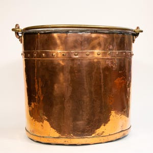 Puede incluir: Un cubo de cobre con asa y borde de latón. El cubo cilíndrico tiene una banda remachada en el centro. El cobre tiene un tono marrón rojizo cálido, con algunas áreas que muestran un acabado más brillante y reflectante. El cubo probablemente se utiliza con fines decorativos o funcionales.