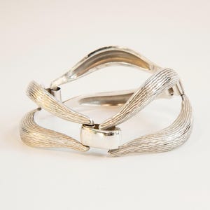 Könnte beinhalten: Ein silbernes Armband mit einem strukturierten, wellenförmigen Design. Das Armband ist offen und hat einen Scharnierverschluss.