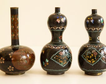 Peut inclure: Trois vases anciens en cloisonné noir avec des motifs floraux et géométriques complexes. Les vases ont une base bulbeuse et un col fin, avec des détails en émail coloré. Le plus grand vase mesure environ 18 cm de haut.