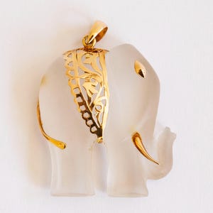 Puede incluir: Un colgante de elefante de vidrio transparente con un diseño de filigrana dorada alrededor de la cabeza y un bucle dorado para colgar.