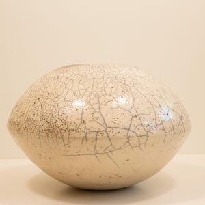 Pode incluir: Um grande vaso de cerâmica branco sujo com um padrão de esmalte rachado. A forma arredondada tem uma superfície salpicada e texturizada com linhas cinza escuro. O vaso é uma peça de arte decorativa.