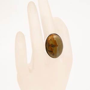 Puede incluir: Anillo ovalado con una piedra preciosa grande y pulida en tonos marrones y verdes. La piedra está engastada en una banda plateada. El diseño del anillo es sencillo, destacando la belleza natural de la piedra.