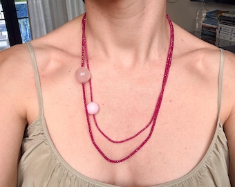 Puede incluir: Un collar largo de doble hebra con pequeñas cuentas facetadas de color rosa intenso. El collar está acentuado con dos esferas lisas, redondas y de color rosa claro de diferentes tamaños, situadas a un lado de las hebras para crear un efecto de capas.
