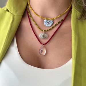 Op de afbeelding: Drie gelaagde kettingen met gedraaide koordkettingen in geel, olijfgroen en donkerrood. De bovenste ketting heeft een witte halfronde steen met een kleine vierkante paarse edelsteen. De middelste heeft een doorschijnende ovale steen en de onderste een heldere ovale hanger met een donkerrood midden.