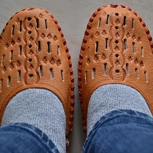 May include: A pair of brown leather shoes with intricate cutouts and red stitching. The shoes are worn with gray socks and blue jeans.