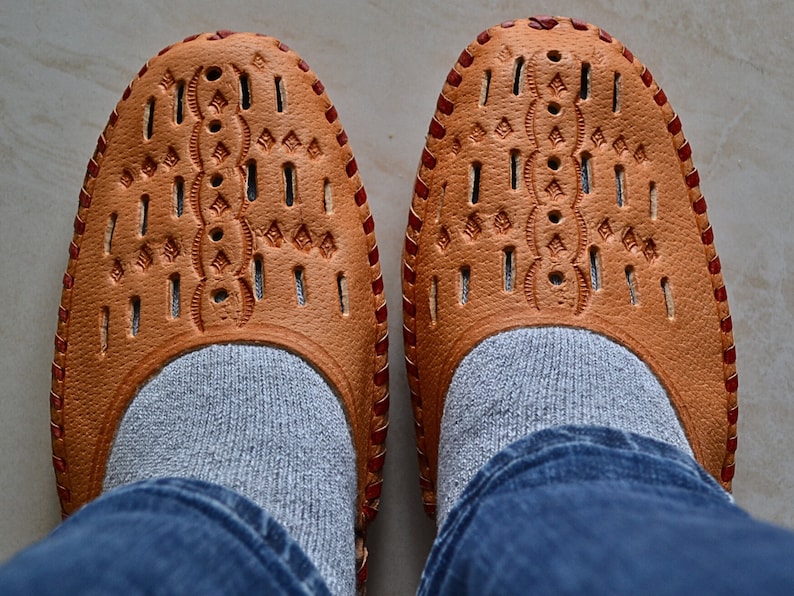 May include: A pair of brown leather shoes with intricate cutouts and red stitching. The shoes are worn with gray socks and blue jeans.