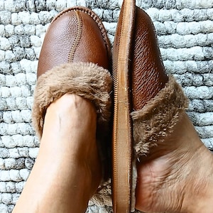May include: Brown leather slippers with faux fur trim. The slippers have a stitched design on the top and a soft, cushioned interior. The image shows the slippers from a top-down perspective, highlighting their cozy and comfortable appearance.