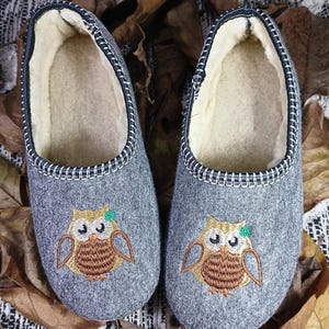 May include: A pair of gray wool slippers with a black trim and an embroidered owl design. The owls are brown with green eyes and a green leaf on their head.