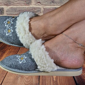 以下が含まれることがあります： 白いフェイクファーのトリムと刺繍された花が付いたグレーのフェルトスリッパ。スリッパはライトグレーのソールです。
