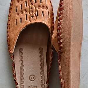 May include: A pair of brown leather slippers with red stitching. The slippers have a decorative pattern on the top and a closed toe.
