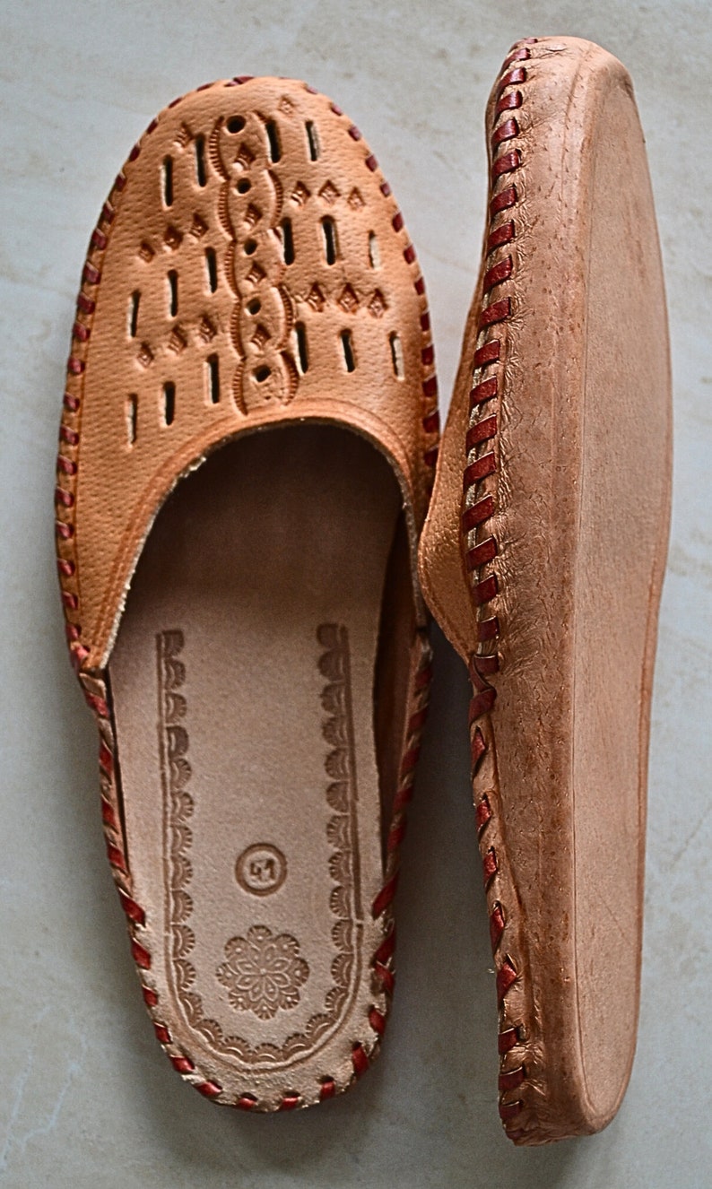 May include: A pair of brown leather slippers with red stitching. The slippers have a decorative pattern on the top and a closed toe.