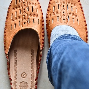 May include: A pair of tan leather slippers with red stitching and a decorative pattern. The slippers have a closed toe and an open heel. The slippers are worn by a person wearing blue jeans.