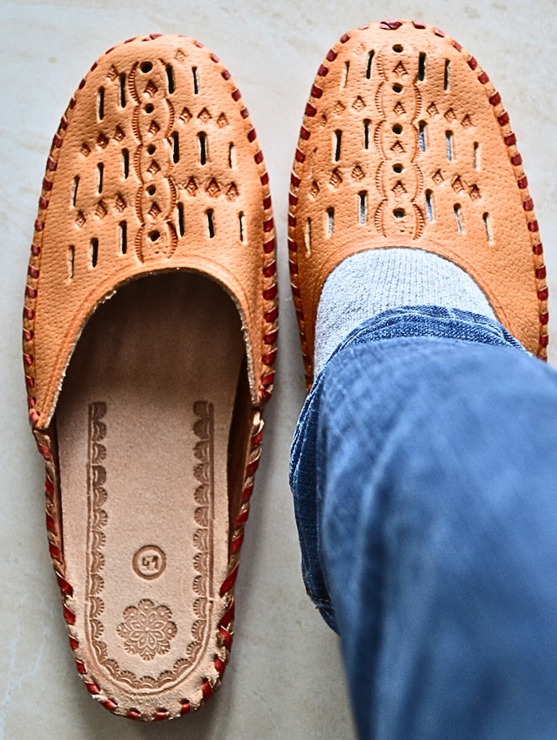 May include: A pair of tan leather slippers with red stitching and a decorative pattern. The slippers have a closed toe and an open heel. The slippers are worn by a person wearing blue jeans.