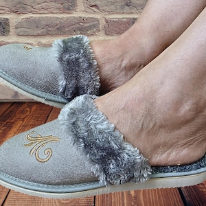 May include: A pair of gray fuzzy slippers with a gold embroidered design. The slippers are worn by a person's feet.