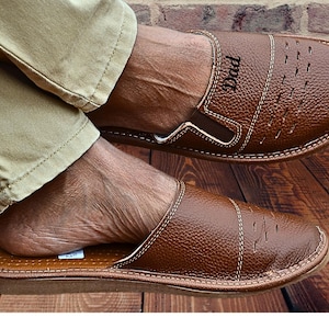 May include: Brown leather slippers with the word "Dad" stitched on the top. The slippers have a slip-on design with decorative stitching and small cut-out details. The image shows the slippers being worn.
