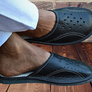 Puede incluir: Un par de zapatillas de cuero negro con costuras blancas y un diseño perforado. Las zapatillas tienen la punta cerrada y un estilo de deslizamiento.