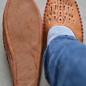 May include: A pair of brown leather slippers with a decorative pattern on the top. One slipper is worn on a foot with blue jeans.