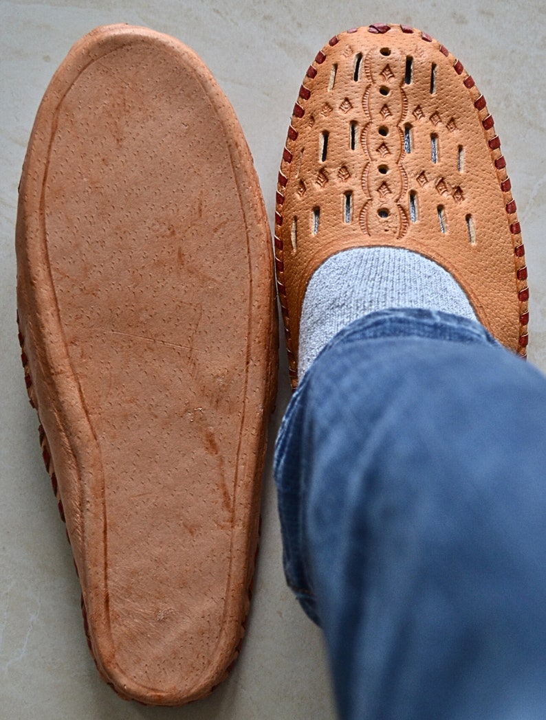 May include: A pair of brown leather slippers with a decorative pattern on the top. One slipper is worn on a foot with blue jeans.
