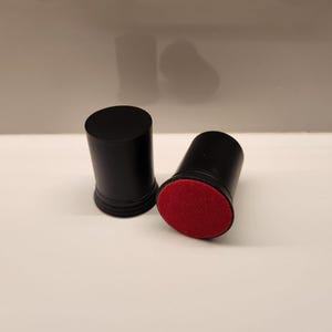 May include: Two black cylindrical bottle stoppers. One stopper has a red felt bottom. The stoppers are on a white surface. The stoppers are made of a solid material.