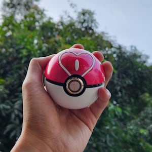Può includere: Una Poké Ball rosa e bianca con un design a cuore sulla parte superiore. La palla ha un anello color oro attorno al centro e una striscia nera che separa le sezioni rosa e bianca. È tenuta in mano su uno sfondo verde sfocato.