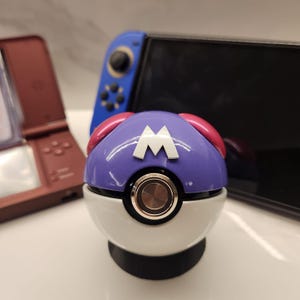 Puede incluir: Un accesorio temático de Poké Ball morado, blanco y rosa con un logotipo "M" blanco. El accesorio tiene un botón metálico y se asienta sobre una base negra. Una Nintendo Switch y una Nintendo 3DS marrón están en el fondo.