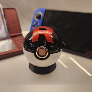 May include: A black and white Poké Ball with a red "R" emblem. The ball has a gold-coloured button and sits on a black base. A Nintendo Switch and a red Nintendo DS are in the background.